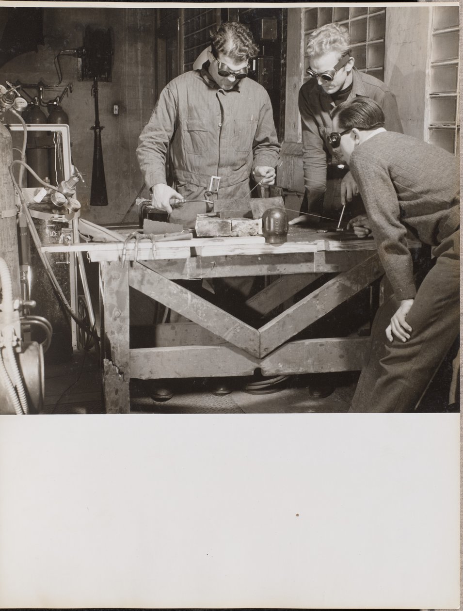 [Trois hommes portant des lunettes de protection soudent une pièce de métal sur une table]. - Arnold Eagle