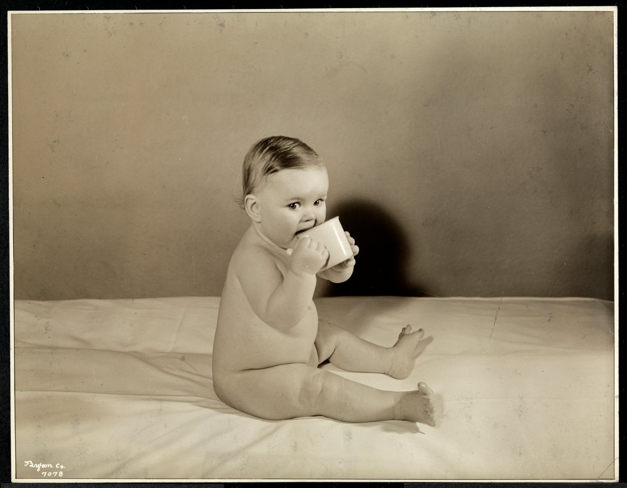 Un bébé assis dans son lit buvant dans une tasse au Maternity Center Association à East River, New York, 1935 - Byron Company