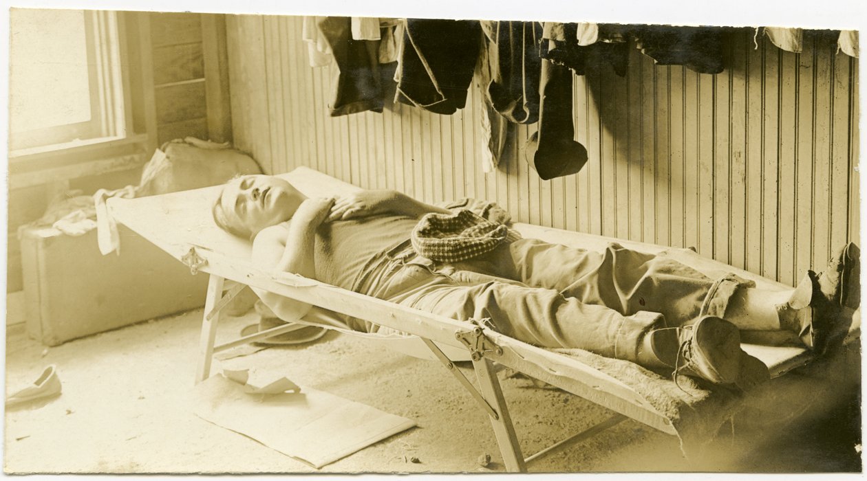 Photographie horizontale sépia montrant un homme dormant sur un lit de camp dans un bâtiment à panneaux de bois. Divers vêtements sont accrochés au mur et des valises sont posées au sol dans le coin de la pièce - Carl Michel