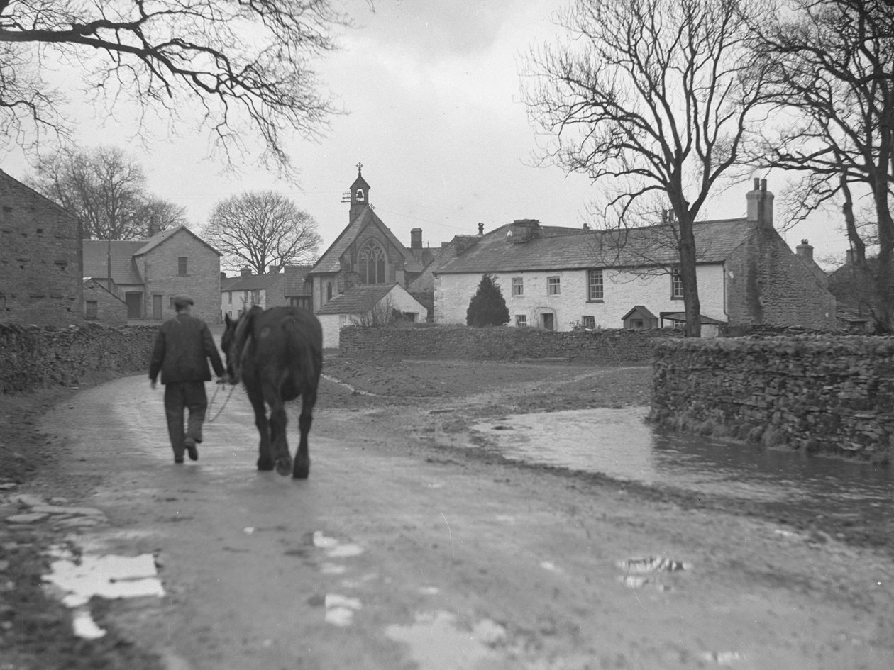 Homme menant son cheval sur la route, loin de la caméra, vers les bâtiments du village et l