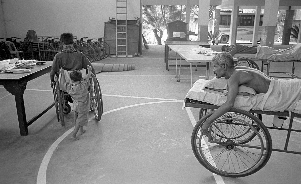 Un homme handicapé en fauteuil roulant est porté par son fils; un autre homme dans la même situation le regarde au Centre de Réhabilitation des Paralysés, CRP à Savar, Dhaka, Bangladesh. 2005. - Unbekannt Unbekannt