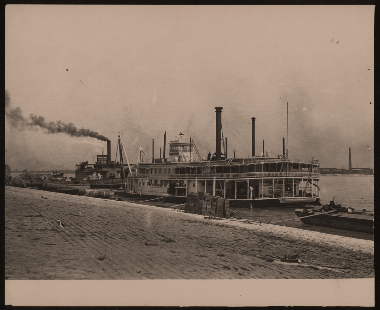 Photographie horizontale en noir et blanc du bateau à vapeur C.R. Suter. Un autre bateau à vapeur est garé derrière et des caisses sont empilées sur le rivage - William G. Swekosky