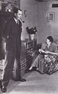 La femme lit, tandis que le mari se réchauffe le dos contre le feu, vers 1950