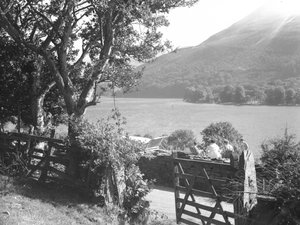 Quatre personnes assises sur un muret de pierres sèches en train de pique-niquer avec vue sur le lac, voiture garée avec coffre ouvert. - Joseph Hardman