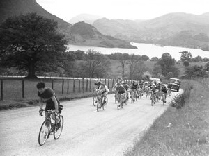 Groupe de cyclistes remontant une route suivis par des voitures, lac et montagnes en arrière-plan
