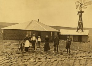 Famille de colons devant leur maison en terre avec un moulin à vent sur le toit du bâtiment adjacent dans le comté de Custer, Nebraska, vers 1885 (photo n/b)