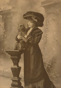 [Sarah Bernhardt debout dans un studio, tenant un petit chien noir sur un piédestal en bois sculpté et orné.] - Theodore C. Marceau