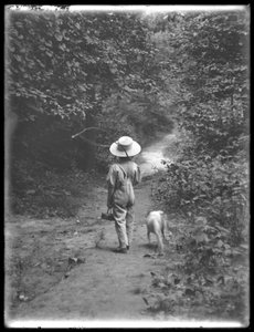 William Gray Hassler en chapeau de paille et salopette, portant une canne à pêche et un panier, marchant sur un chemin dans les bois avec un petit chien (Bessie), 20 août 1911