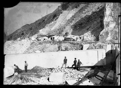 Trois hommes observent le travail de plusieurs excavatrices dans les Alpes apuanes. Derrière eux se trouve un énorme bloc de marbre déjà taillé et prêt à être transporté dans la vallée - A. Corsini