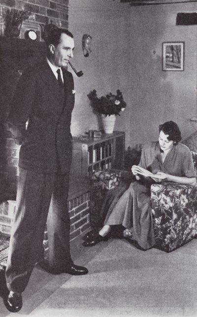 La femme lit, tandis que le mari se réchauffe le dos contre le feu, vers 1950 - English Photographer