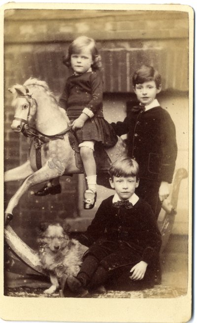 Jeune fille sur un cheval à bascule avec un garçon debout et un garçon assis avec un chien - English School