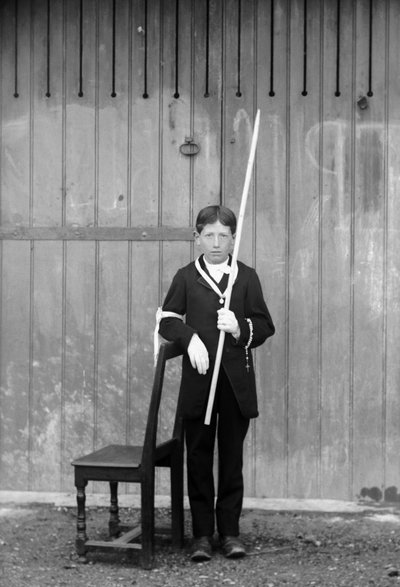 Premier communiant, en costume blanc et gants, avec médailles, chapelet, manchette en dentelle blanche et tenant une très longue bougie, près de Bourmont en Haute-Marne, vers 1880. Photographie. Collection Ducos. - French Photographer