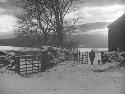 Homme et chien traversant le portail en direction de la caméra dans la neige avec Thirlmere derrière. - Joseph Hardman