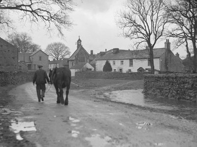Homme menant son cheval sur la route, loin de la caméra, vers les bâtiments du village et l