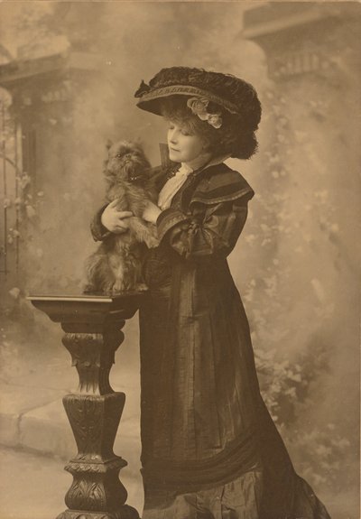 [Sarah Bernhardt debout dans un studio, tenant un petit chien noir sur un piédestal en bois sculpté et orné.] - Theodore C. Marceau