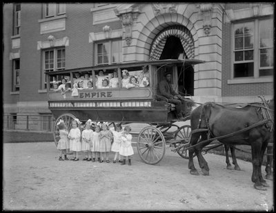 Petites filles posant dans et devant un chariot tiré par des chevaux, devant un bâtiment non identifié, probablement l