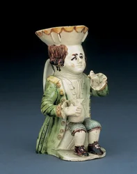 Cruche Toby buveur, son chapeau avec bord festonné brun et jaune, ses cheveux bruns incisés en queue et avec une rosette blanche assis avec une cruche balustre sur son genou, c.1770