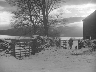 Homme et chien traversant le portail en direction de la caméra dans la neige avec Thirlmere derrière.