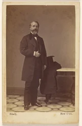 [Homme barbu non identifié debout, un haut-de-forme et un manteau sur une table voisine]