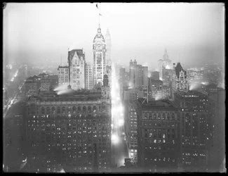 Vue nocturne brumeuse en plongée du bas de Manhattan vers le nord, avec le Singer Building, le Woolworth Building et le Municipal Building visibles, vers 1913