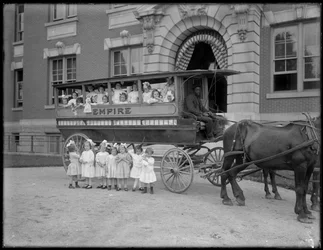 Petites filles posant dans et devant un chariot tiré par des chevaux, devant un bâtiment non identifié, probablement l