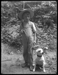 William Gray Hassler en chapeau de paille et salopette, portant une canne à pêche et un panier, posé dans les bois avec un petit chien (Bessie), 20 août 1911